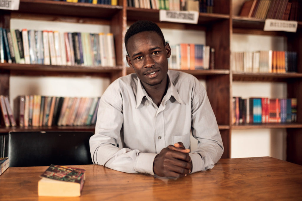 Portrait of Evans Kiprotich sat a table with a bookshelf behind him